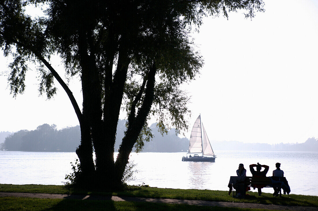 Three people sitting on a park bench on the island Frauenchiemsee, Fraueninsel on Lake Chiemsee, Bavaria, Germany
