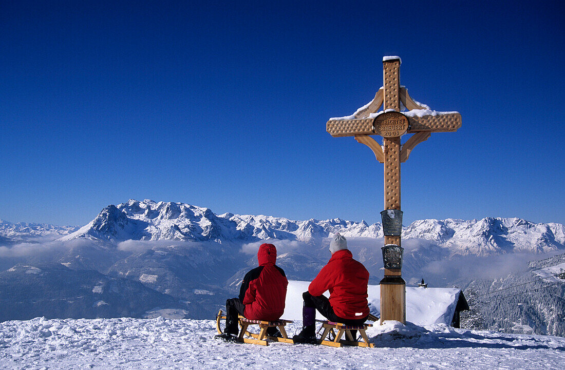 Two persons sledging at cross of Bischlinghöhe, view to Hochkönig range, skiing area Werfenweng, Salzburg, Austria