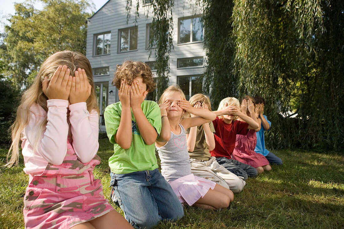 Kinder hocken nebeneinander im Gras und halten sich die Augen zu, Kindergeburtstag