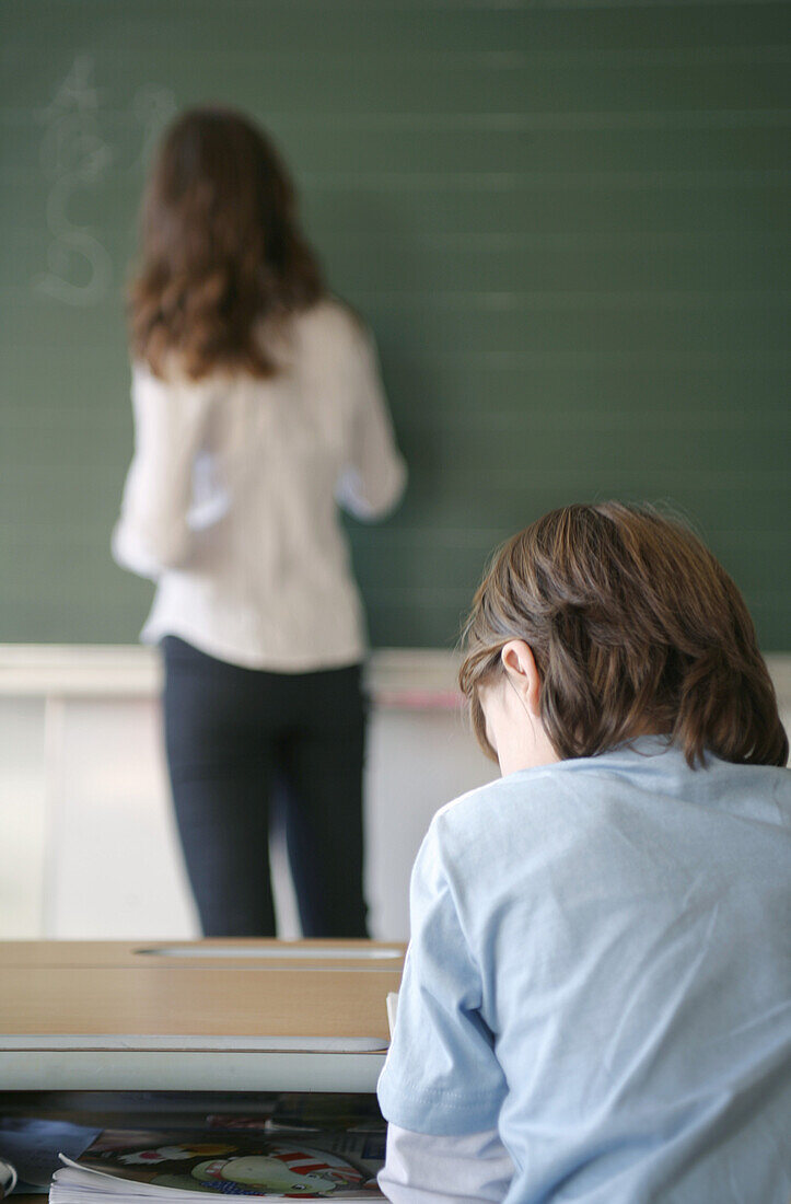Boy sitting in classroom, teacher standing at blackboard