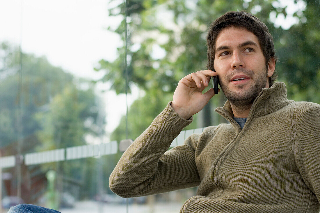 Junger Mann sitzt in einer Bushaltestelle und telefoniert mit einem Handy