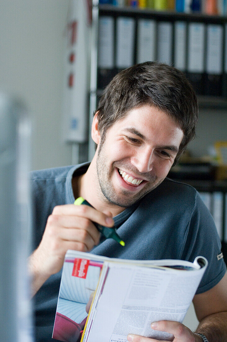 Lächelnder junger Mann liest in einer Zeitschrift, hält Textmarker in der Hand