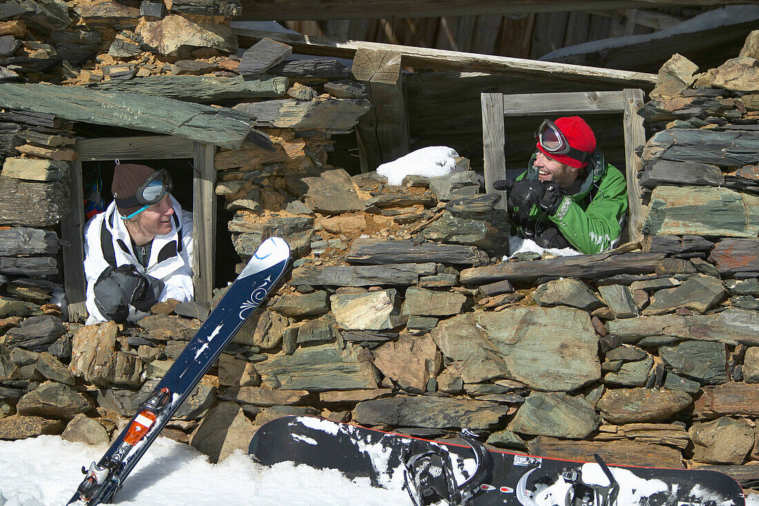 Two snowboarder looking through windows of derelict cabin, Falkertsee, Carinthia, Austria