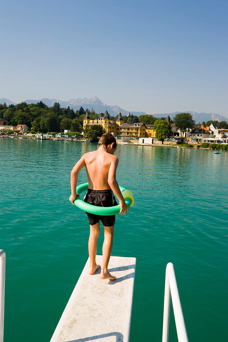 Boy with floating tyre standing on diving platform, Hotel Schloss Velden in background, Worthersee, Velden, Carinthia, Austria