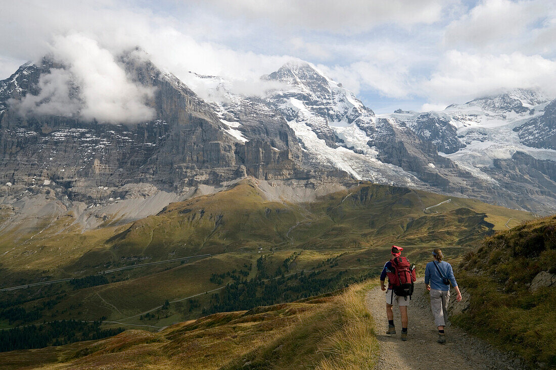 Couple hiking along Männlichen Panoramaweg (panorama hiking path), view to Kleine Scheidegg (2061 m), Eiger (3970 m), Mönch (4107 m) and Jungfrau (4158 m) in background, Grindelwald, Bernese Oberland (highlands), Canton of Bern, Switzerland