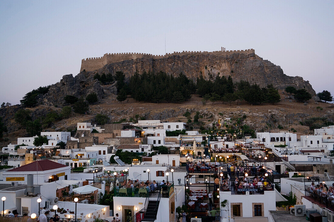 View over illuminated town in the evening to Acropolis, people sitting on terraces of restaurants, Lindos, Rhodes, Greece