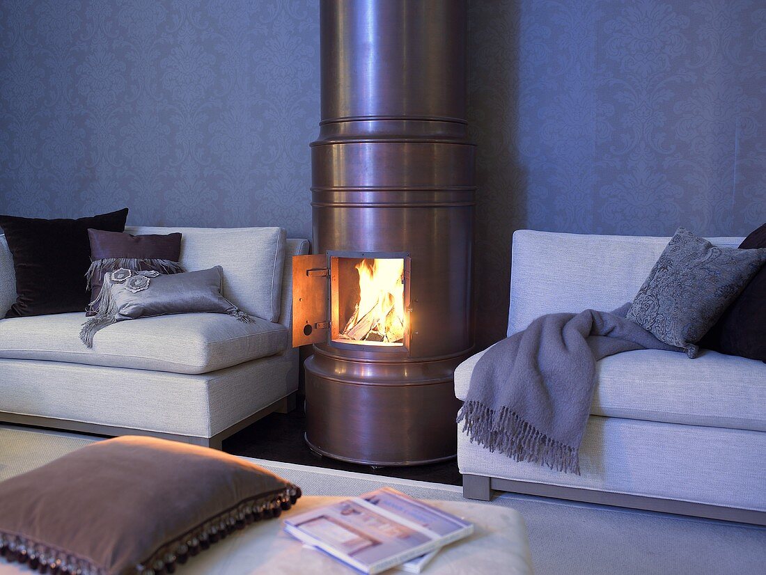 A copper coloured wood burning stove with a open door and view of the fire next to a sofa strewn with pillows and throws
