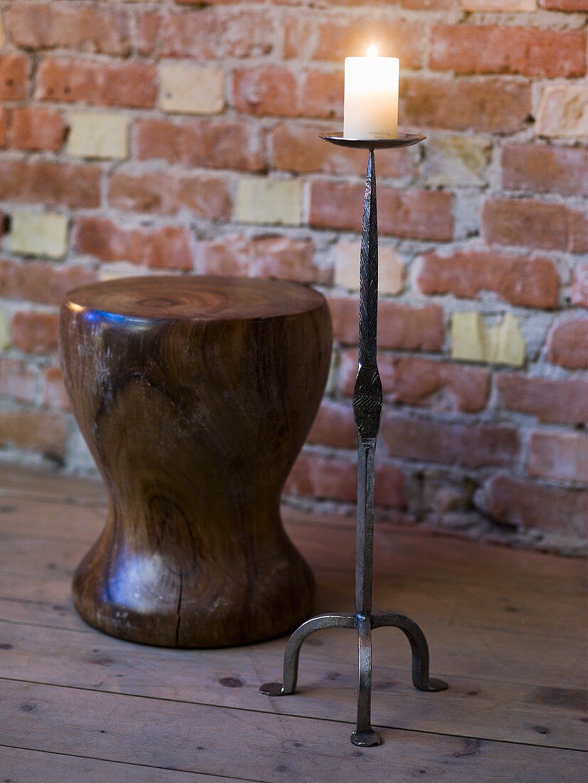 Floor candle stand with burning candle and wooden stool in front of a brick wall