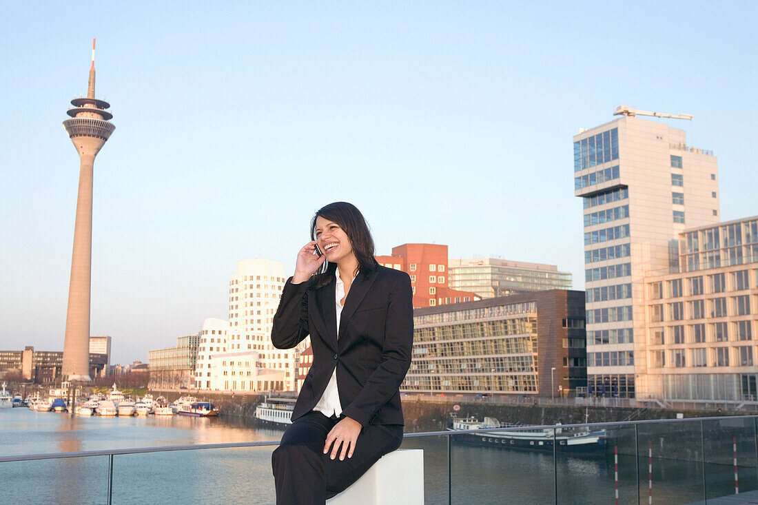 Young business woman talking on mobile phone in front of the city skyline, television tower, Zollhof, Media Harbour, architecture of Frank O.Gehry, Düsseldorf, state capital of NRW, North-Rhine-Westphalia, Germany