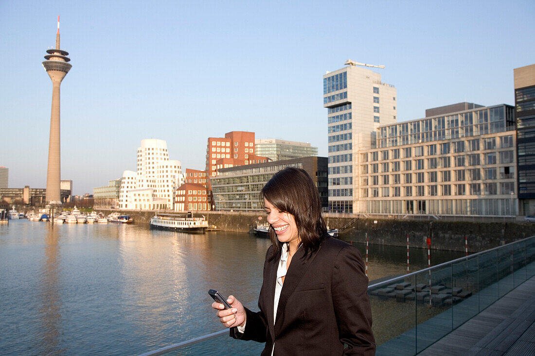 Young business woman writing an SMS, in front of the city skyline, Media Harbour, Düsseldorf with television tower and Zollhof, architecture of Frank O.Gehry, state capital of NRW, North-Rhine-Westphalia, Germany