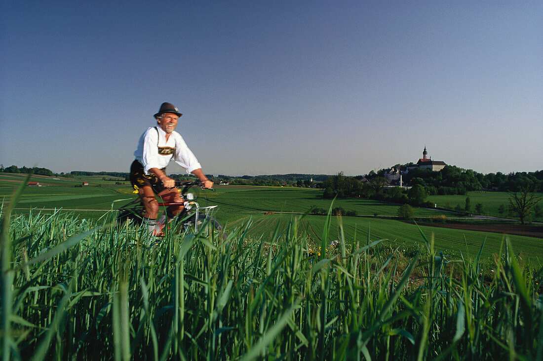 Man wearing traditional costume riding a bicycle, Andechs Abbey in background, Bavaria, Germany