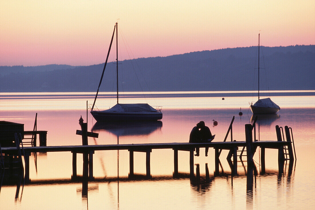 Paar sitzt auf Bootssteg am Starnberger See in der Abenddämmerung, Bayern, Deutschland