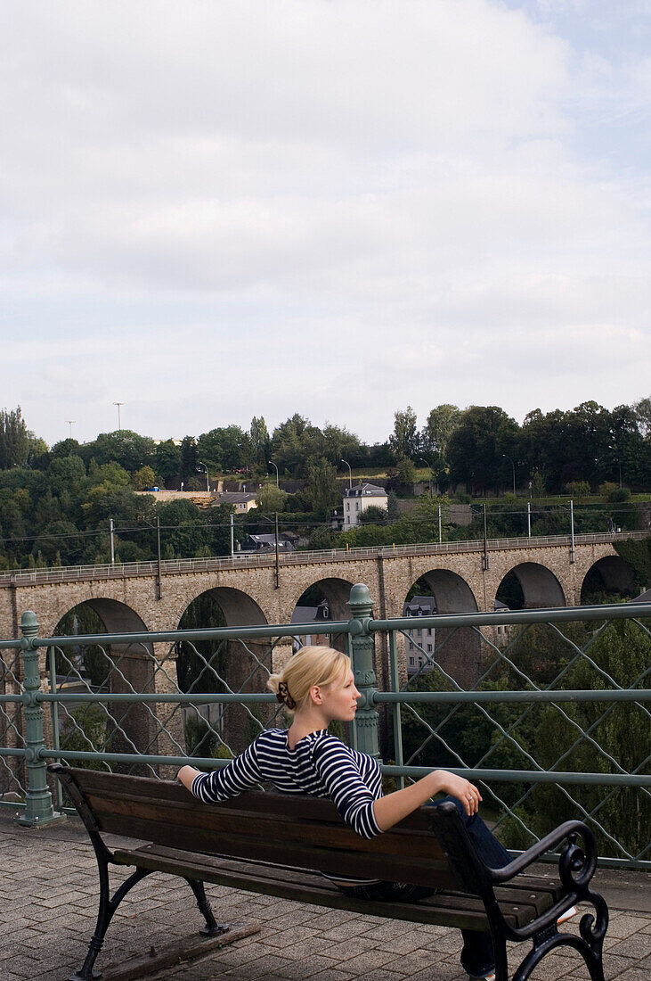 Frau sitzt auf einer Bank und betrachtet die gegenüberliegende Brücke, Luxemburg