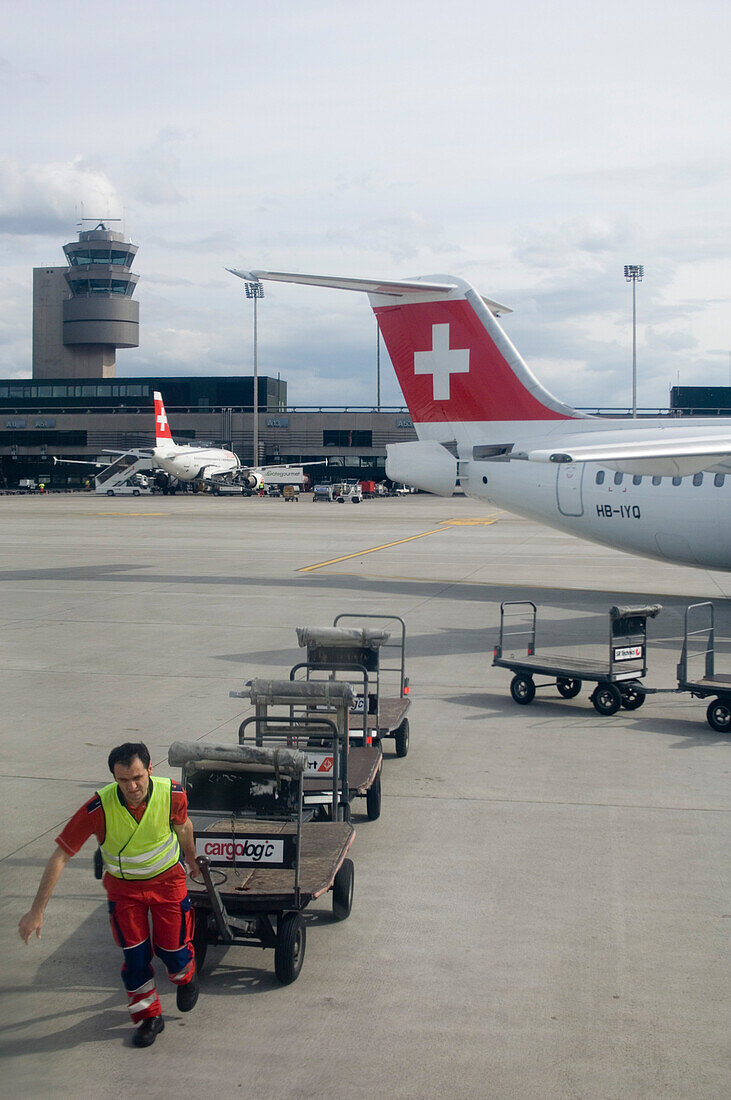 Ein Mann zieht leere Gepäckwagen über eine Landebahn des Flughafens, Zürich, Schweiz