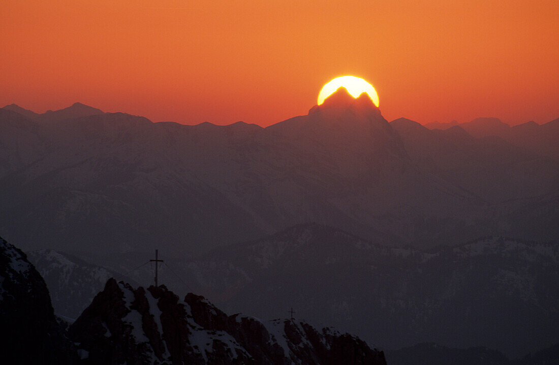 Sonnenscheibe hinter dem Guffert mit Felsgipfeln mit Gipfelkreuz im Vordergrund, Wilder Kaiser, Tirol, Österreich