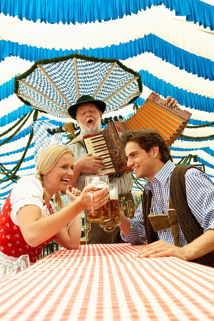 Paar stößt mit Bier im Bierzelt an, Mann im Hintergrund spielt Akkordeon
