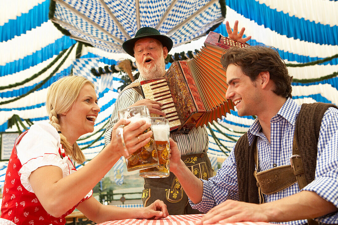 Paar stößt mit Bier im Bierzelt an, Mann im Hintergrund spielt Akkordeon