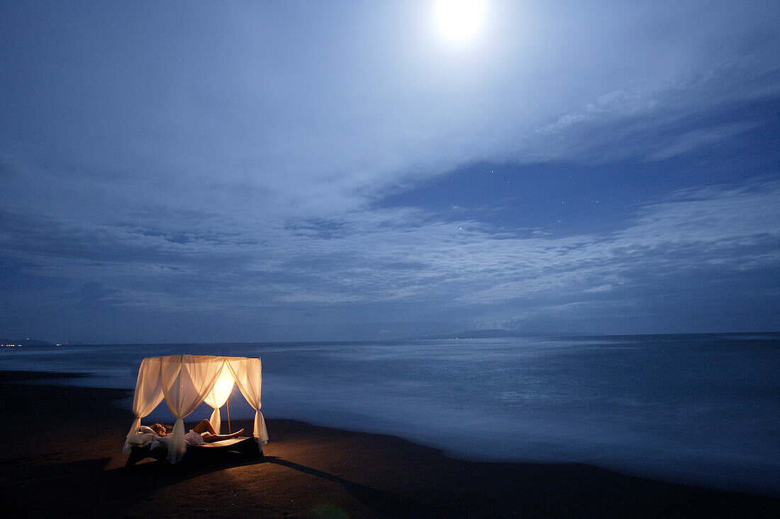 A young woman lying in a four poster bed on the beach in the moonlight, near Uluwatu, Bali, Indonesia