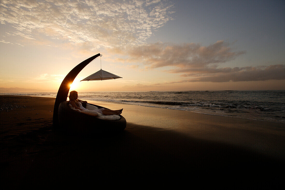 Eine junge Frau entspannt sich am Strand bei Sonnenuntergang, nahe Uluwatu, Bali, Indonesien