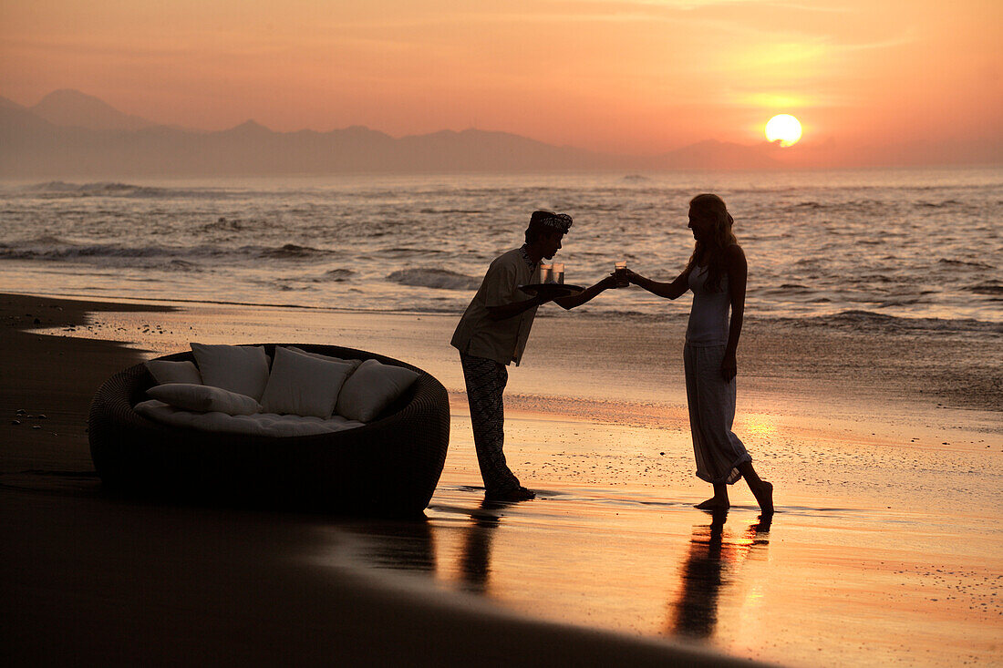 A young woman being offered a drink from a waiter on the beach at sunset, near Uluwatu, Bali, Indonesia