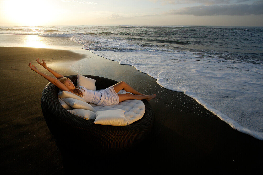 Eine junge Frau entspannt sich am Strand bei Sonnenuntergang, nahe Uluwatu, Bali, Indonesien