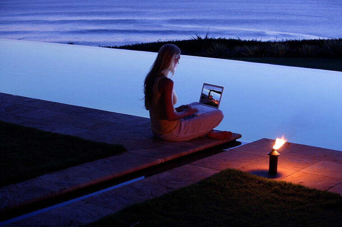 Young woman sitting with laptop at the edge of a swimming pool, looking at photographs, near Uluwatu, Bali, Indonesia
