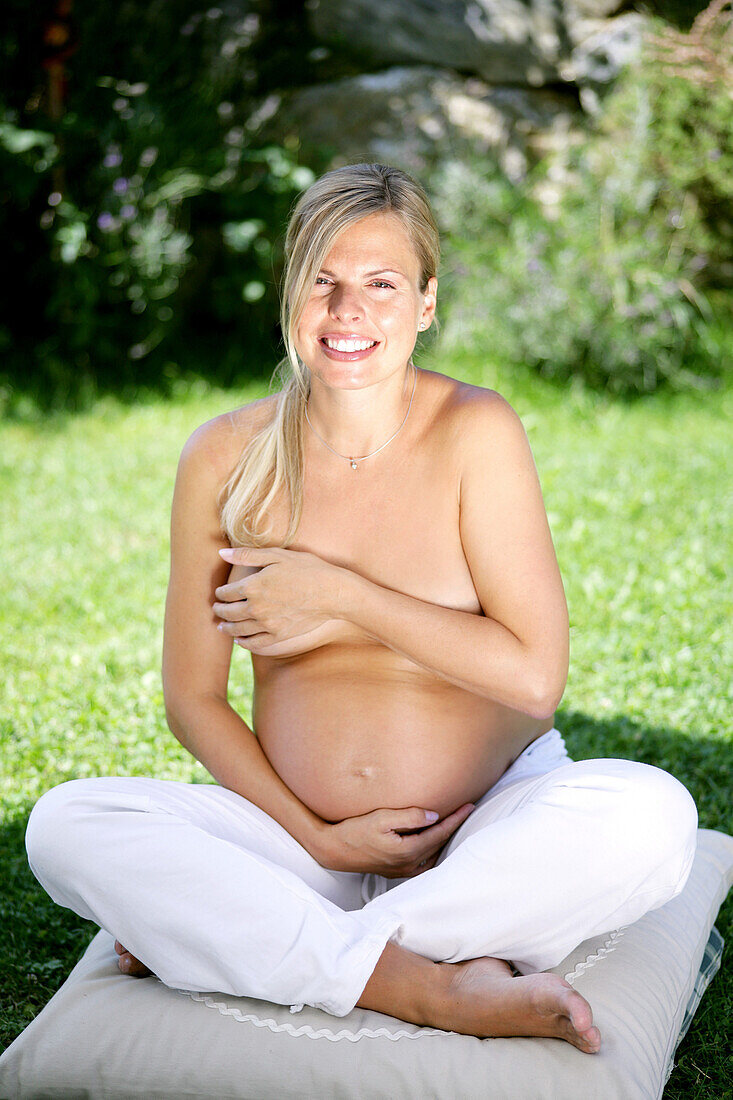 Schwangere Frau sitzt mit freiem Oberkörper im Garten, lächelt in die Kamera, Steiermark, Österreich