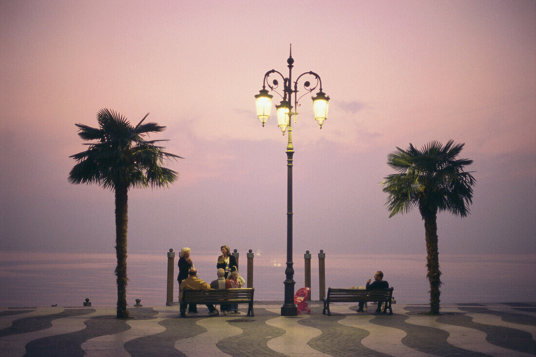 Menschen sitzen abends auf einer Bank der Uferpromenade am Gardasee, Lazise, Provinz Verona, Veneto, Italien
