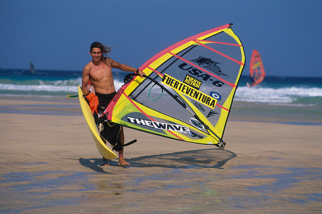 Ein muskulöser Windsurfer trägt Brett und Segel über den Strand Playa Barca, Playa de Sotavento, Jandía Halbinsel, Fuerteventura, Islas Canarias, Kanarische Inseln, Spanien