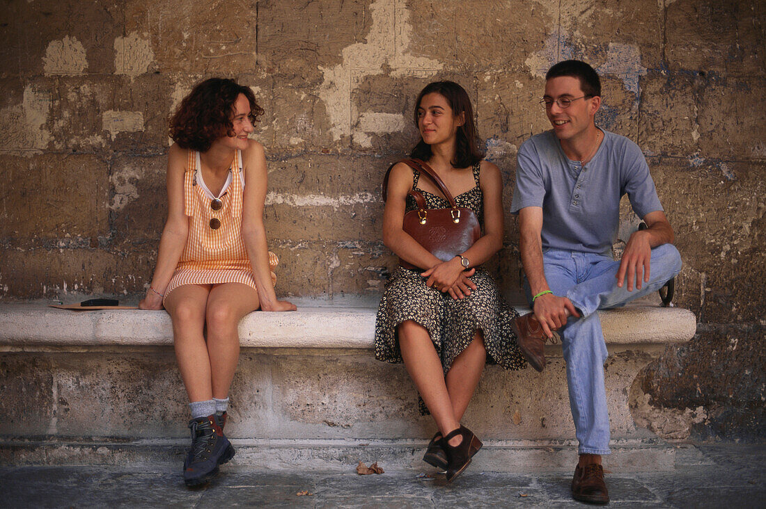 Two young women and a young man sitting on a stone bench in the courtyard of the Colegio de Santa Cruz, Valladolid, Castilla-Leon, Northern Spain