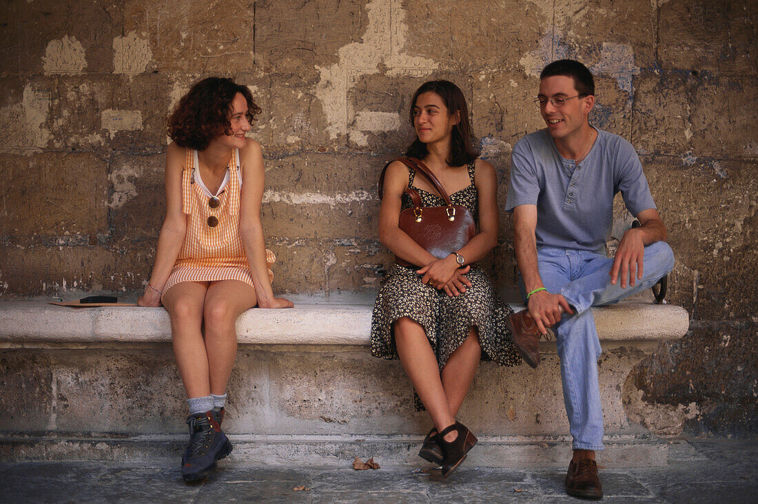 Zwei junge Frauen und ein junger Mann sitzen auf einer Steinbank im Innenhof des Colegio de Santa Cruz, Valladolid, Altkastilien, Castilla-León, Spanien