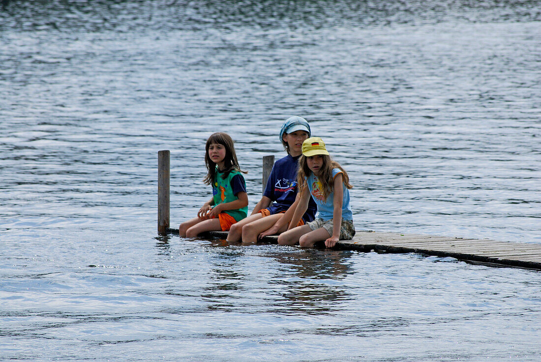 three girls at landing stage with legs in water, lake Staffelsee, Upper Bavaria, Bavaria, Germany