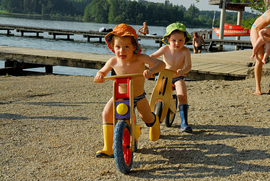 zwei Jungen mit Sonnenhut auf Laufrad, Strandbad am Hartsee, Chiemgau, Oberbayern, Bayern, Deutschland