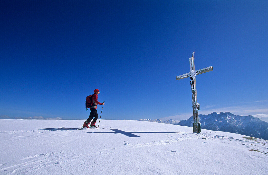 Crosscountry skiier arriving at rime ice covered cross on summit of Fellhorn, view to Loferer Steinberge, Chiemgau range, Chiemgau, Upper Bavaria, Bavaria, Germany