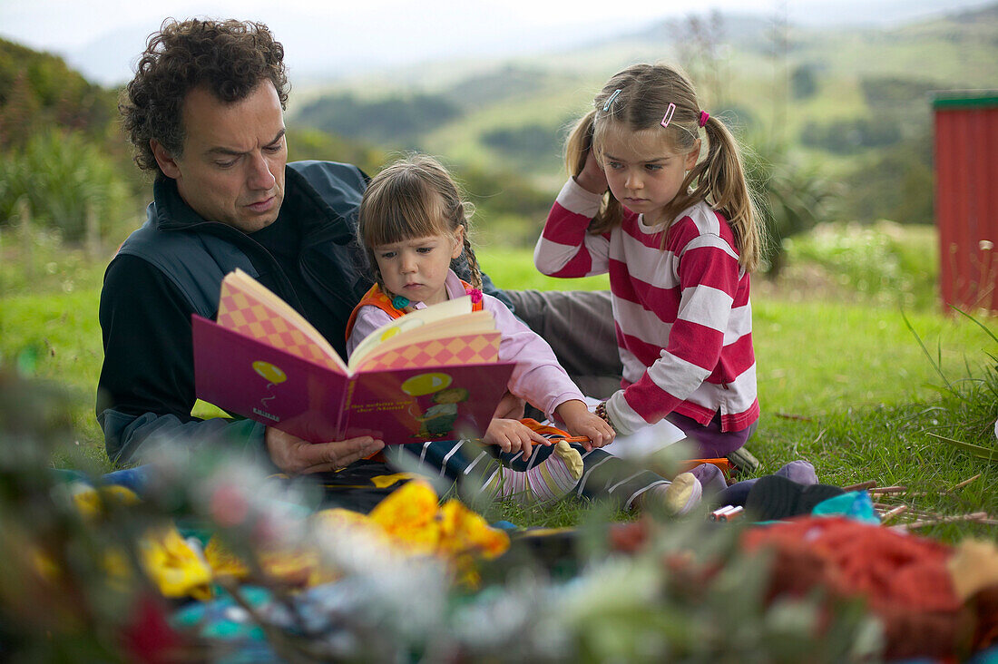 Father and children reading picture book,  Okopako Lodge, near Opononi, at Hokianga Harbour, Northland, North Island, New Zealand