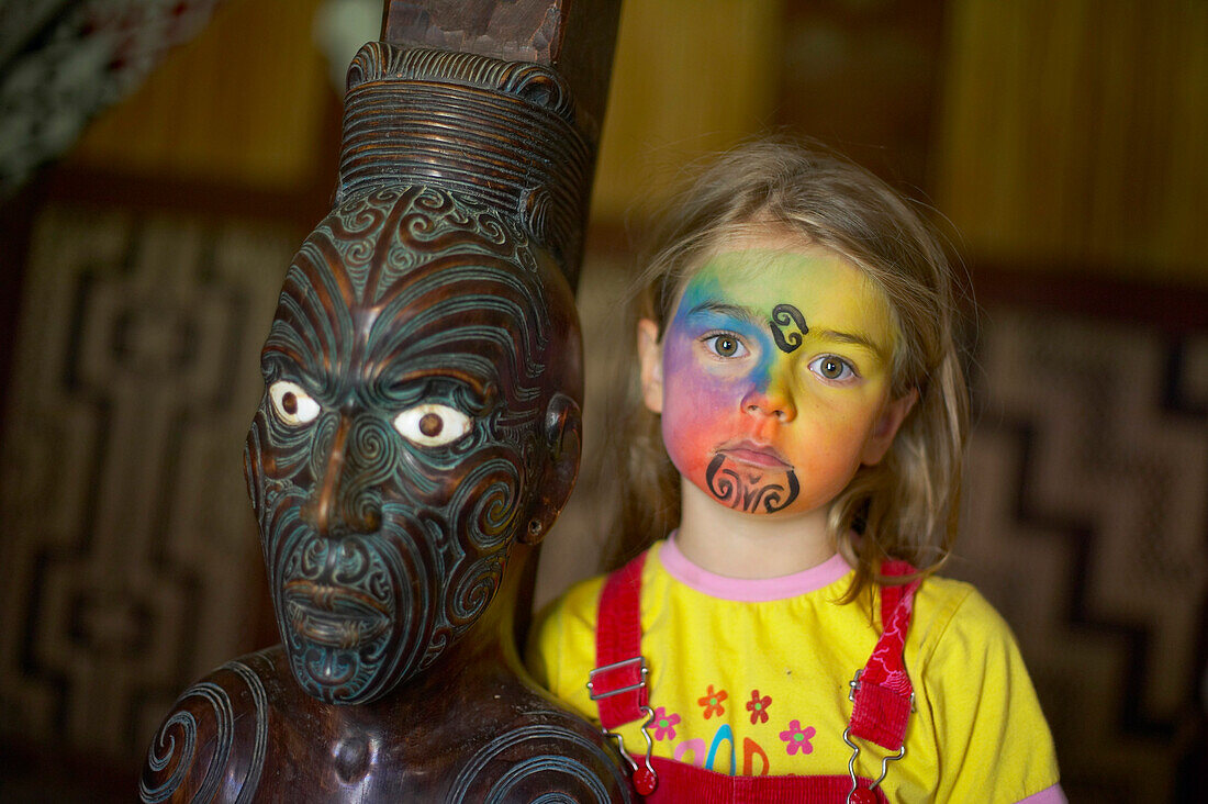 Mädchen mit Facepainting, nachempfundenes Moko (Maori Gesichtstatoo), geschnitzte Maori Figur in Versammlungshaus (Marae),  Okains Bay, Bank's Peninsula, Ostküste, Südinsel, Neuseeland