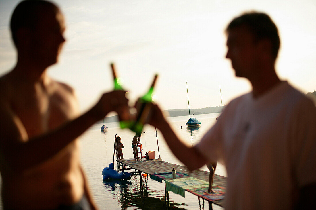 Two men drinking bottles of beer, children playing on jetty in background, Lake Woerthsee, Bavaria, Germany, MR