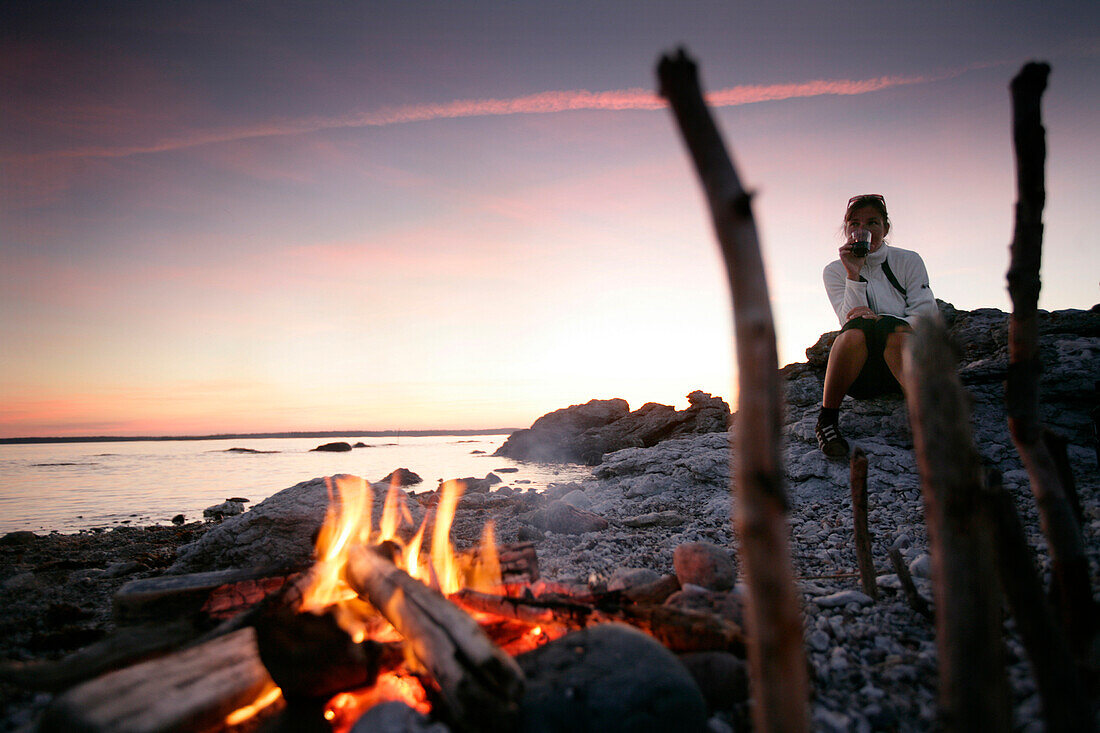 Frau trinkt Wein beim Lagerfeuer am Strand, Sonnenuntergang, Sysne, Gotland, Schweden