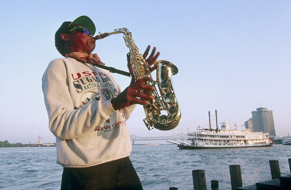 Jazz musician playing saxo on Mississipi bank and steamboat Natchez in background. Louisiana, USA