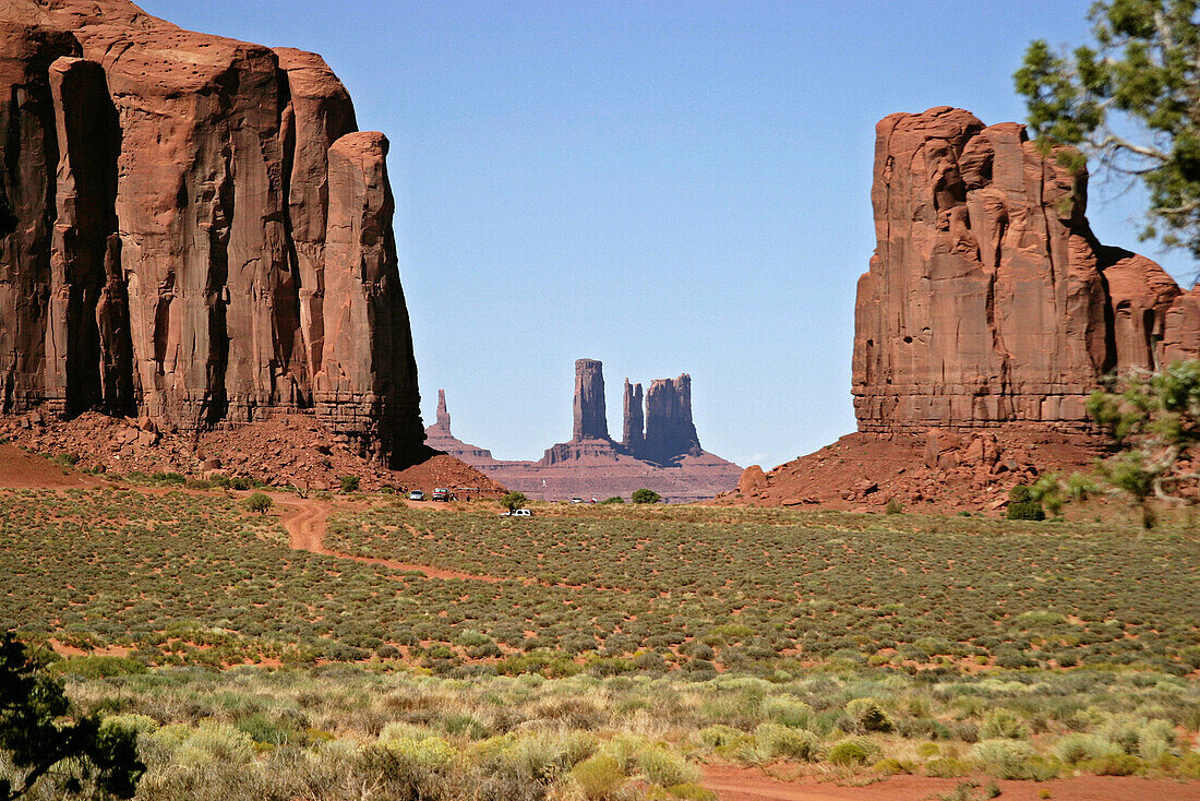 Arid green plants in foreground with two huge red rock formations framing distant red rock formation with vehicles in frame to lend scale. Monument Valley Navajo Tribal Park. Arizona-Utah, USA