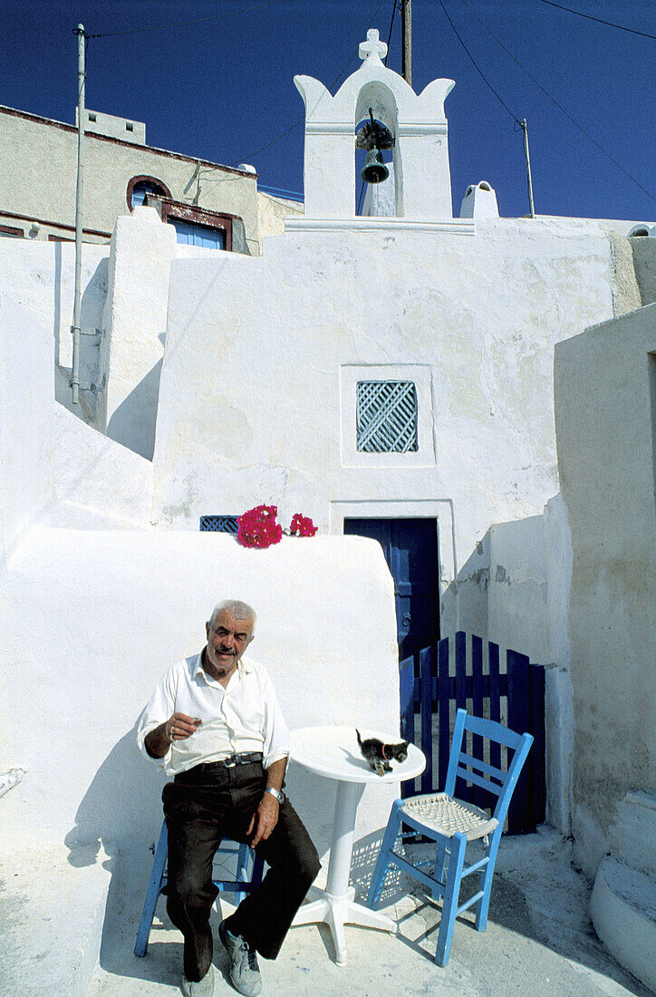 Small cafe owner sitted at his only table on terrace.White washed houses and church belfry at back. Ia village. Santorini (Cyclades). Greece