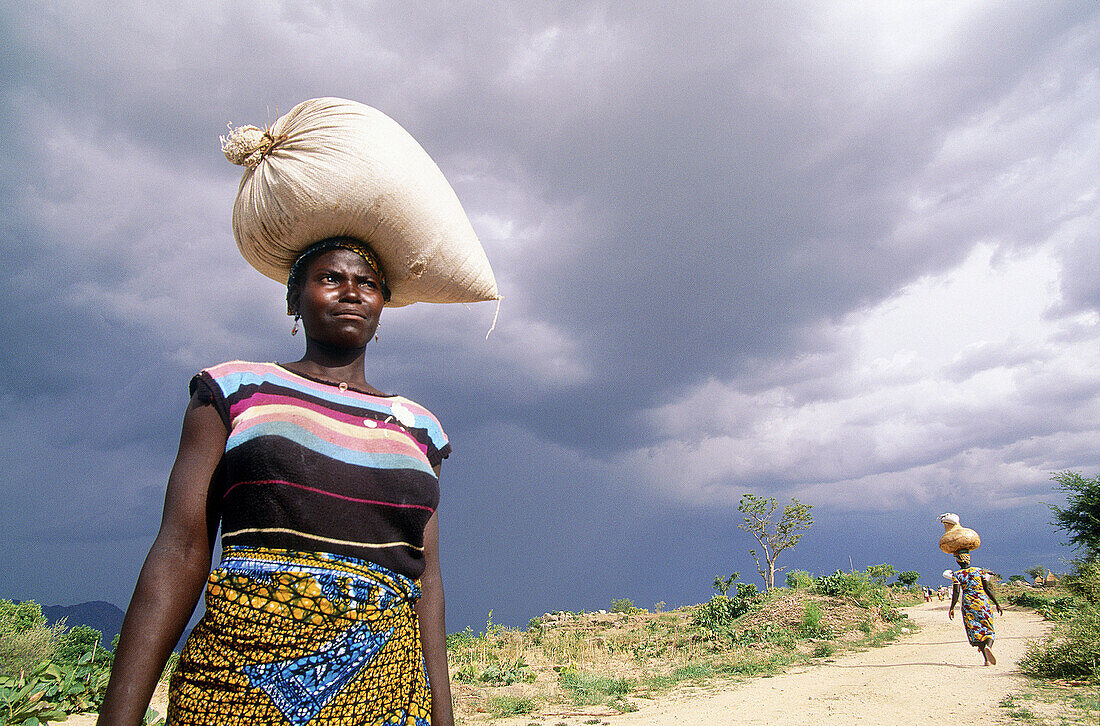 Women carrying heavy loads in their heads under stormy clouds on the trail to Tourou village (Goudours tribe from Nigeria) Mandara Mounts. Cameroon