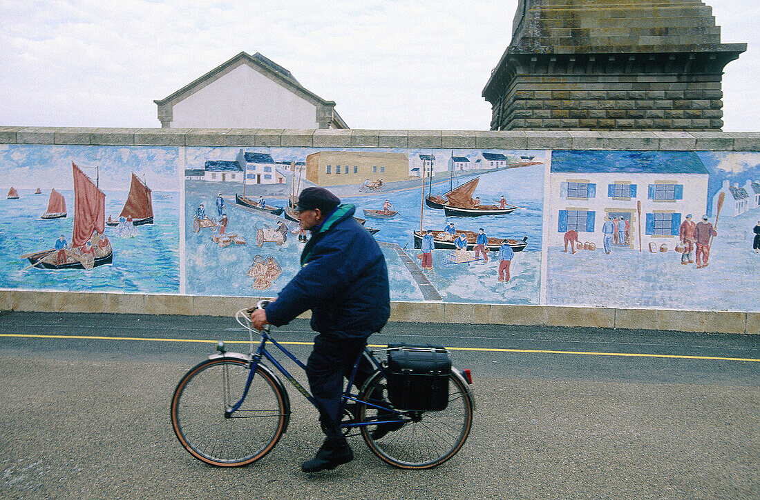 Retired marin cycling by a road with a mural painting wall and Eckmül lighthouse in background. Penmarc h. Finistere. Brittany. France
