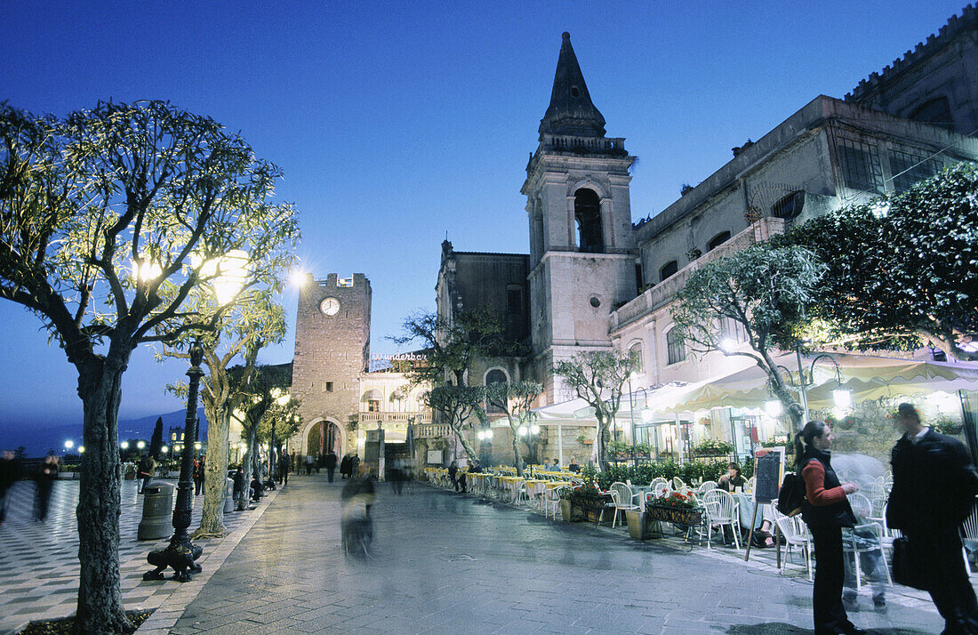 Italy. Sicily. Taormina. Twilight at lit Piazza IX Aprile with clock tower and gate (Porta di mezzo) in distance and tourists, locals, cafes.