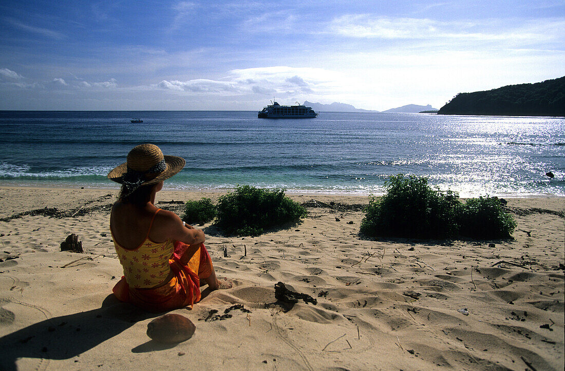 Frau sitzt am Strand und betrachtet den Aussicht, Kreuzfahrtschiff MV Reef Escape im Hintergrund, Navadra Island, Mamanuca Gruppe, Fidschi, Südsee