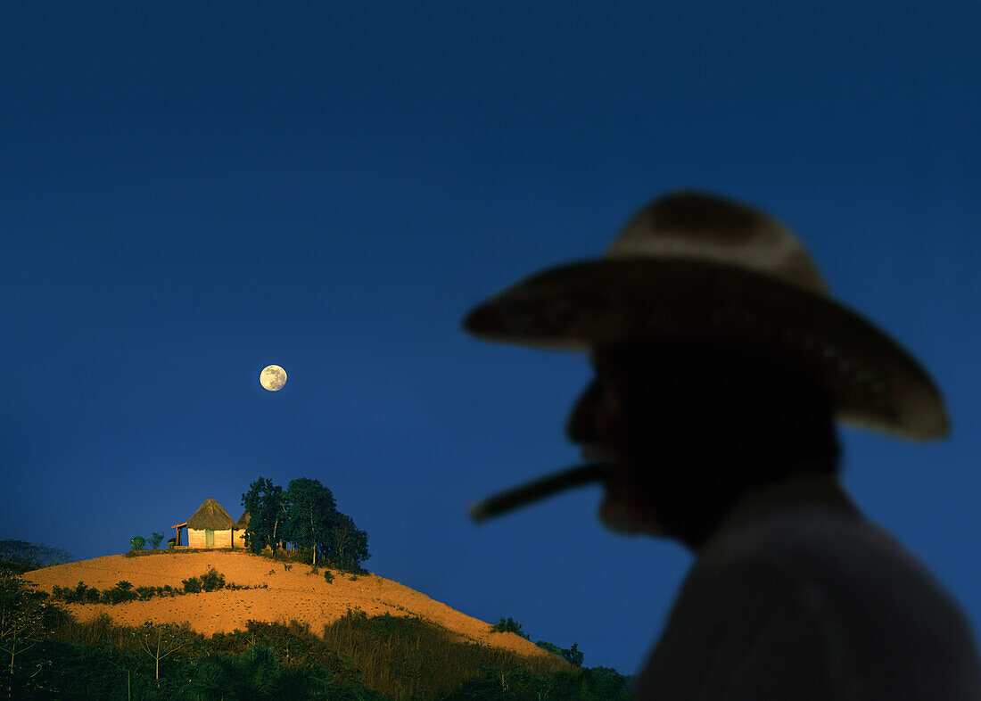 Bauer mit Hut raucht eine Zigarre, Mondaufgang über Tabak Felder im Hintergrund, Vinales Valley, Pinar del Rio, Cuba, Karibik