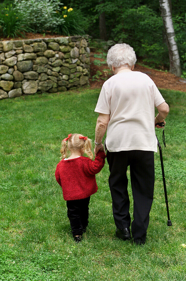 Senior woman, grandmother, walking with cane across backyard lawn with her 3 year old granddaughter.
