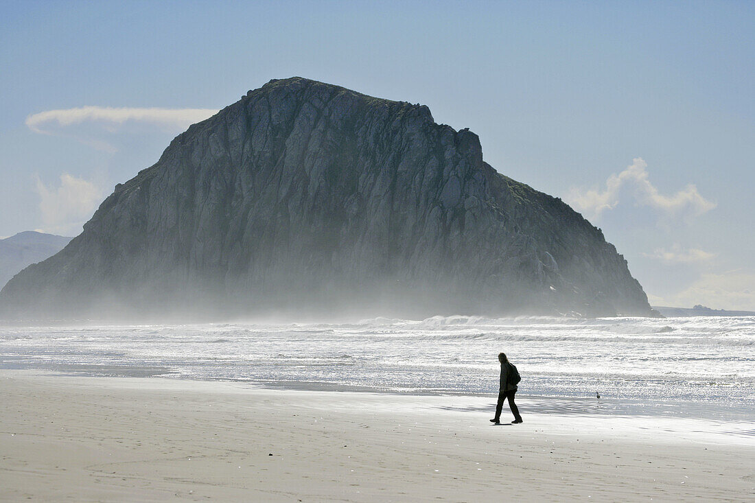 Woman walking through the salt air, sea spray on beach in front of Morro Rock, Morro Bay, CA