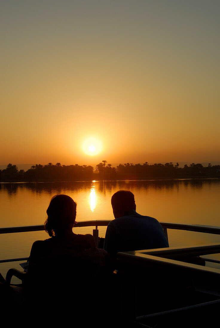 cruise on the Nile, couple on upper deck, sunset above palm trees at western bank, Nile between Luxor and Dendera, Egypt, Africa