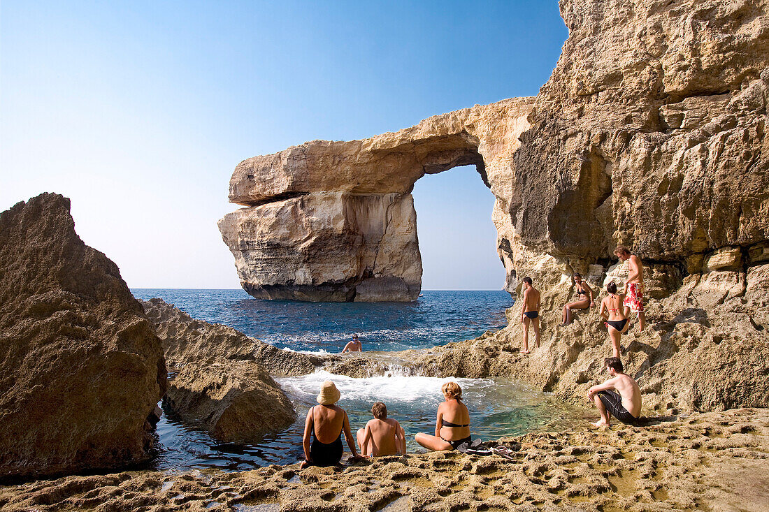 Menschen sitzen am Meer auf Felsen, Felsbogen im Hintergrund, Gozo, Malta, Europa